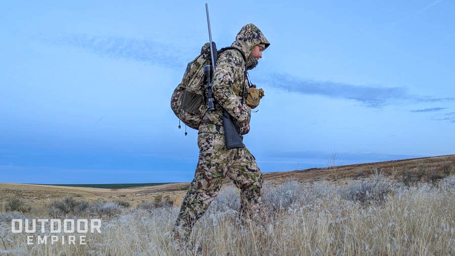 Hunter walking through dry grass with rifle on shoulder and wearing sitka camouflage clothing